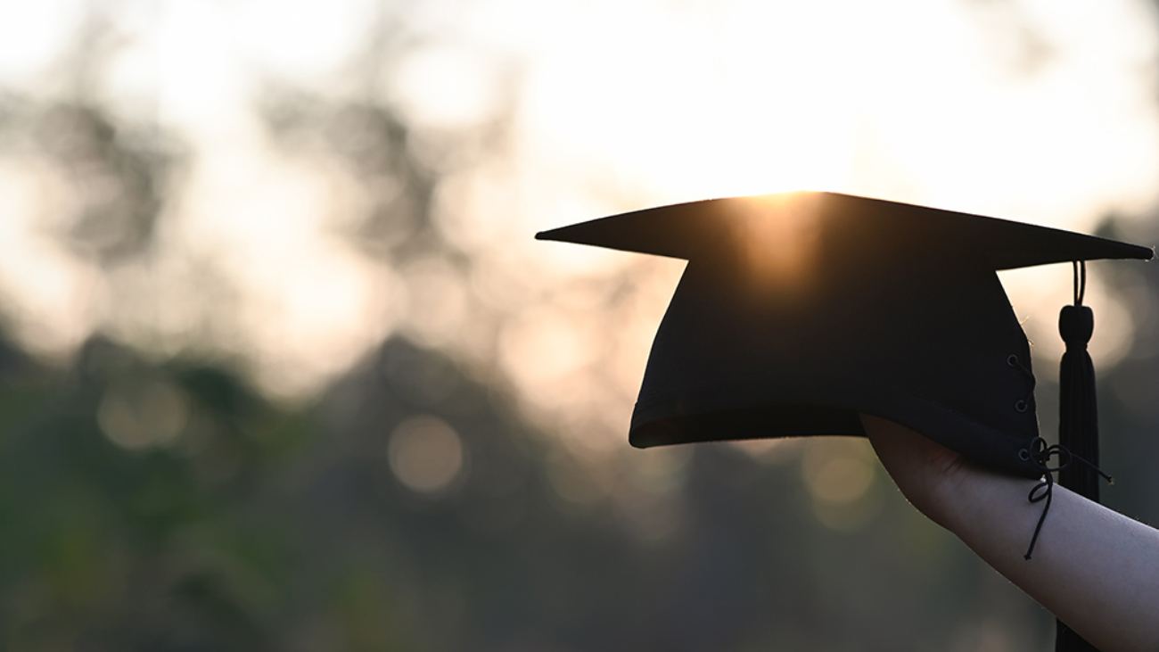 Cropped image of university student hand holding a graduation hat in hand over outdoors with sunset as background.