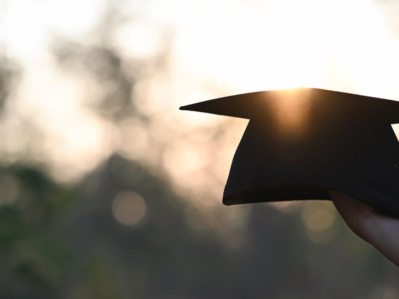 Cropped image of university student hand holding a graduation hat in hand over outdoors with sunset as background.