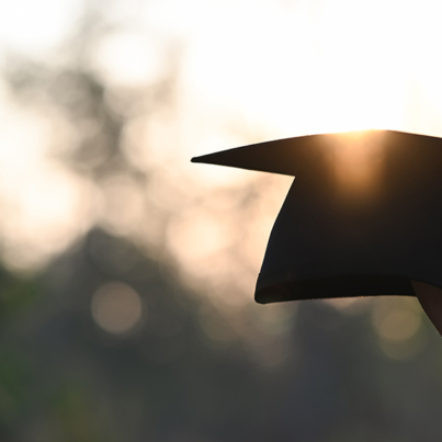 Cropped image of university student hand holding a graduation hat in hand over outdoors with sunset as background.