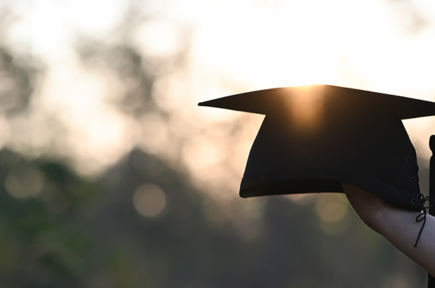 Cropped image of university student hand holding a graduation hat in hand over outdoors with sunset as background.