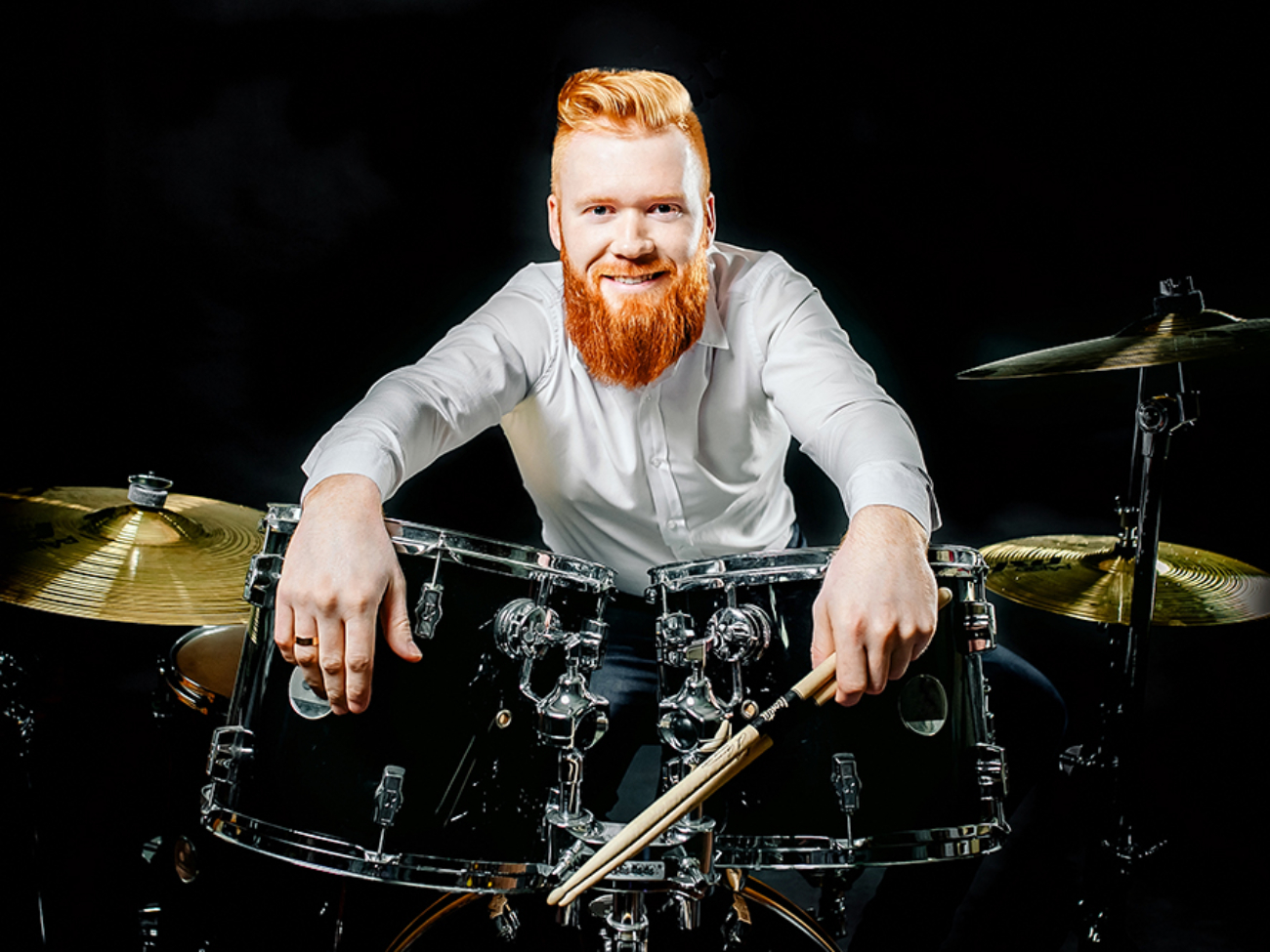 Portrait of a red-haired emotional man playing drums and cymbals and holding a stick. isolated on a dark background