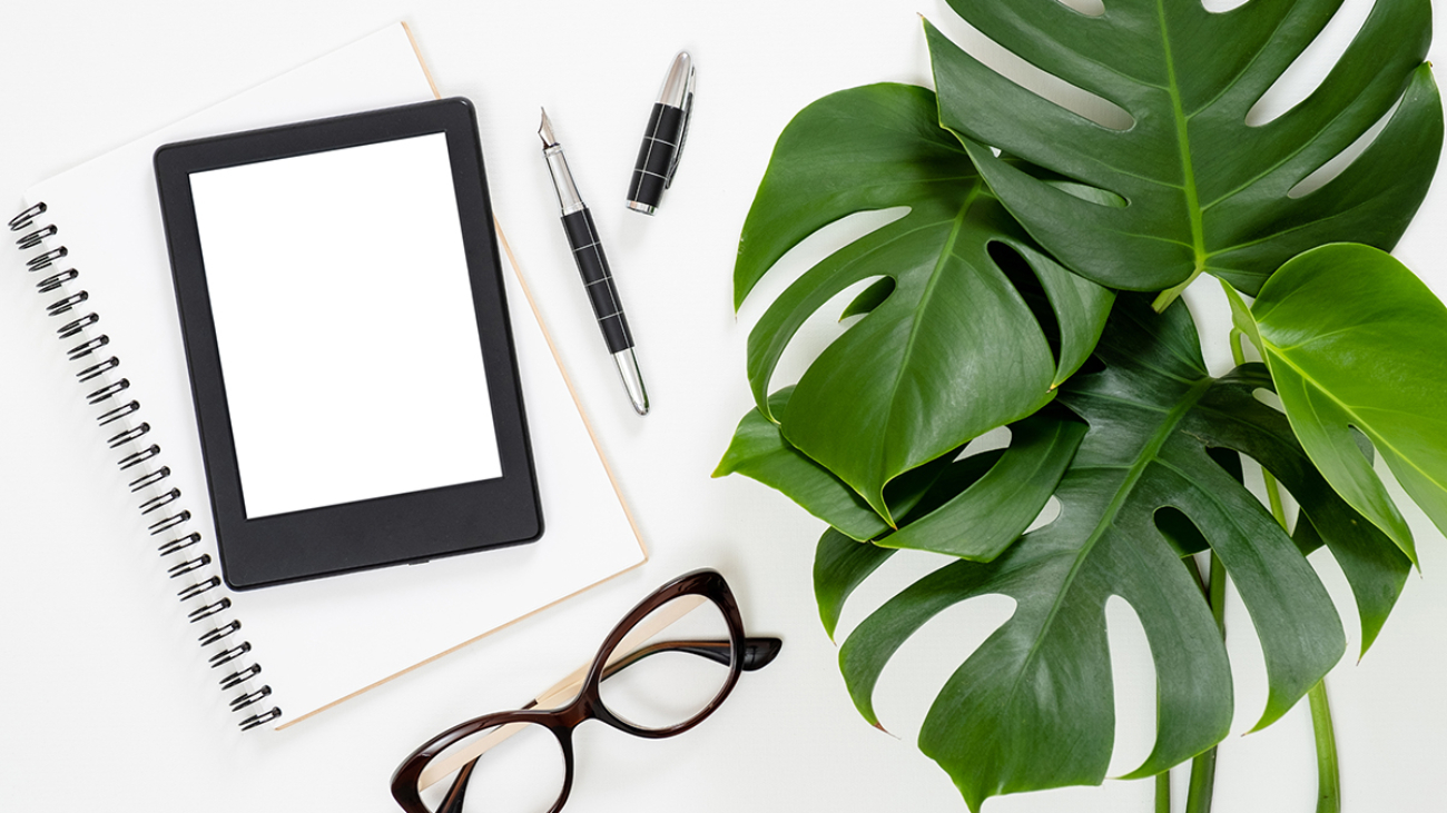 Flat lay tropical jungle Monstera leaves, paper notebook, e-book reader, glasses on white background. Top view feminine diary, stationery and electronic device on home office desk.