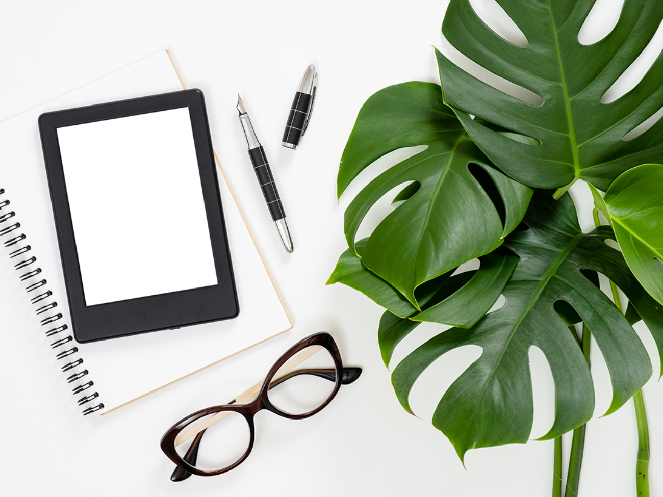 Flat lay tropical jungle Monstera leaves, paper notebook, e-book reader, glasses on white background. Top view feminine diary, stationery and electronic device on home office desk.