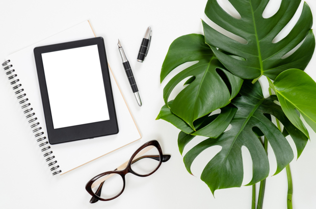 Flat lay tropical jungle Monstera leaves, paper notebook, e-book reader, glasses on white background. Top view feminine diary, stationery and electronic device on home office desk.